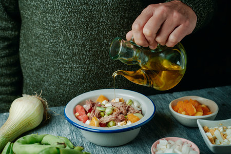 a man dresses with some olive oil a broad bean salad typical of andalusia, made with tomato, onion, boiled egg, tuna and orange, on a rustic gray wooden tableの写真素材