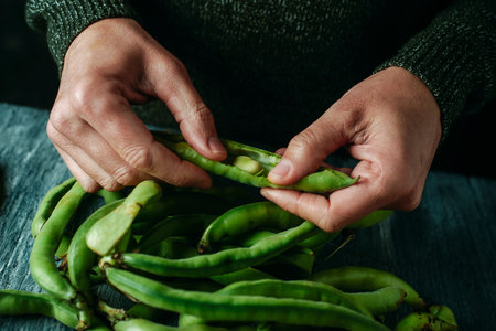 closeup of a man shelling some broad beans at a rustic gray wooden table full of broad bean pods