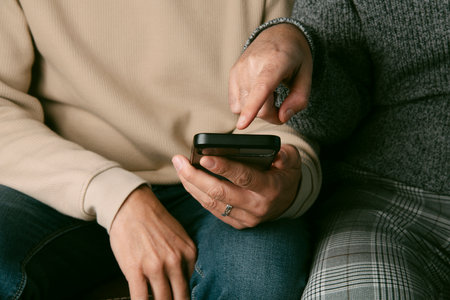 closeup of two men wearing casual clothes, sitting side by side on a brown sofa, and one of them shows something in his smartphone to his partnerの写真素材