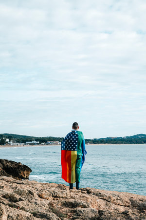a person wrapped in a rainbow US flag stands next to the ocean, with some blank space in the skyの写真素材