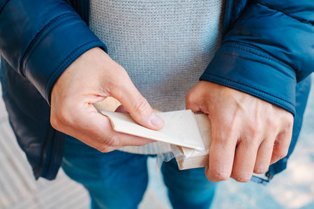 a man, wearing jeans, a gray sweater and a blue jacket, takes a recycled paper tissue out from a tissue packet outdoorsの写真素材