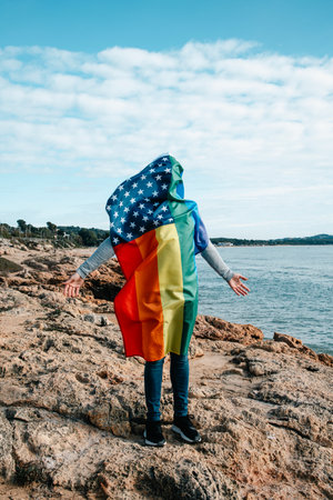 a LGBTIQ person covered with a rainbow US flag stand in front of the ocean, with open armsの写真素材