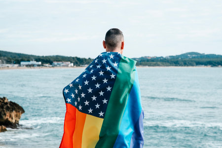 closeup of a man wrapped in a rainbow american flag standing next to the ocean, looking out at itの写真素材