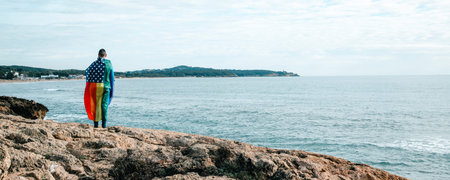 a man wrapped in a rainbow US flag standing next to the ocean, looking out at it, in a panoramic...