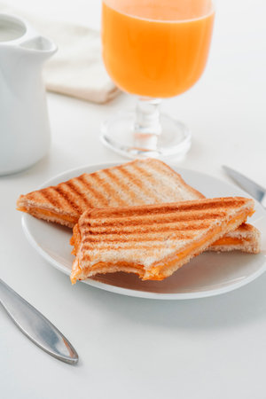 a white ceramic plate with a grilled ham and cheese sandwich, on a white table next to a glass of natural orange juice and a ceramic creamer with milkの写真素材