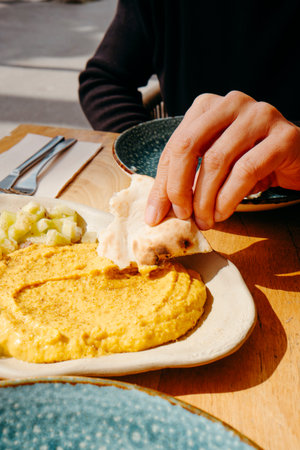 a man dips a piece of naan bread on a curry hummus served on a ceramic plate, sitting at an outdoors...