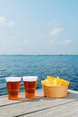 two plastic cups filled with beer and a paper container with potato chips on the old wooden boards of a dock by the waterの写真素材