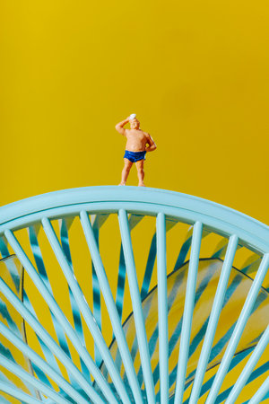 a miniature man in blue swim trunks wipes his forehead while standing on top of a turquoise fan against a vibrant yellow background in a playful and surreal summer sceneの写真素材