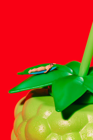 a miniature surfer lays on the leafy lid of a plastic bottle shaped like a green blackberry, on a red backgroundの写真素材