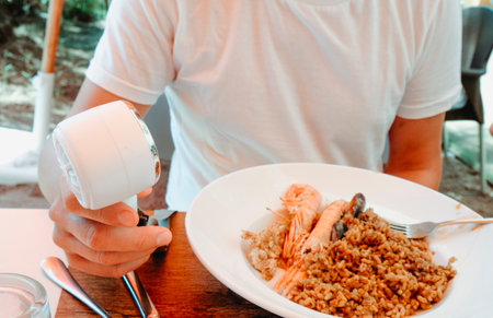 seafood rice with prawns and mussels fills the plate of a man who sits at a wooden table outdoors holding a battery-operated fan to cool down in the summer heatの写真素材