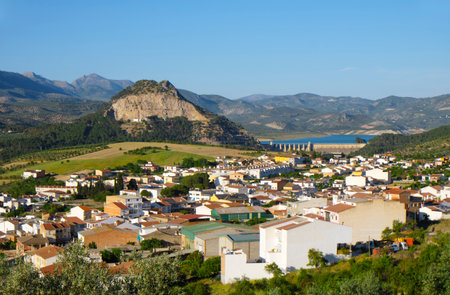 Cuevas de San Marcos, in the Spanish province of Malaga, extends across the valley beneath a rocky hill, with the Iznajar dam and shimmering reservoir visible beyond the fields and rooftopsの写真素材