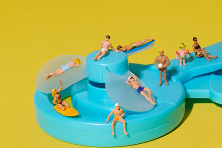 a group of miniature beachgoers including a surfer, swimmers and children is arranged across a blue hand-powered fan with turquoise structure and translucent bladesの写真素材