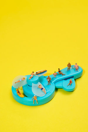 the whole surface of a blue hand-powered fan is covered with miniature people in swimsuits, some sitting, lying or standing, with a solid yellow backgroundの写真素材