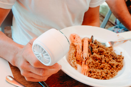a man in a white t-shirt cools himself with a small handheld fan while sitting at an outdoor restaurant table in front of a plate of seafood rice on a warm summer dayの写真素材