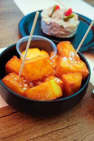 spanish patatas bravas served with a spicy dip appear in the foreground while a portion of ensaladilla rusa, a cold potato salad with vegetables and mayonnaise, is visible on a black plate behindの写真素材