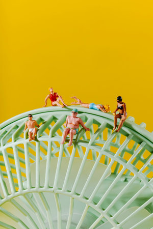 five miniature people in swimwear sit or lie on top of a green household fan with radial spokes, placed in front of a vibrant yellow backdropの写真素材