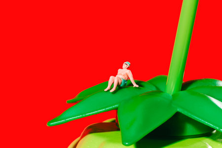 a miniature man in swim trunks and cap sits back with arms extended on a plastic green leaf placed on top of a bottle shaped like a green blackberry, against a flat red backgroundの写真素材