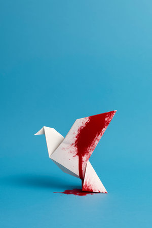 a white origami dove stands on a blue surface with its wing stained by a red blood splatter, creating a striking metaphor of peace wounded and fragile in violenceの素材