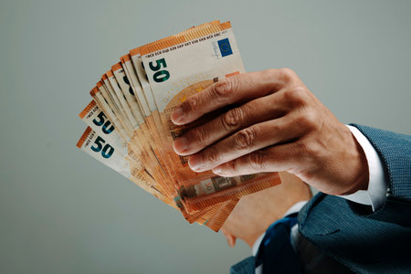 seen from below, a businessman in a blue suit, shirt, and tie holds euro banknotes in his hands while standing indoors, suggesting power or financial controlの写真素材