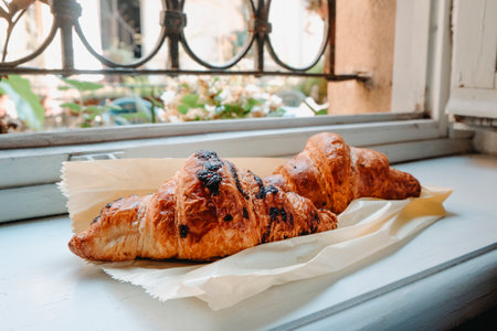 two croissants rest on parchment paper over a white windowsill, one in focus with dark chocolate spots, the other behind, with blurred flowers outsideの写真素材