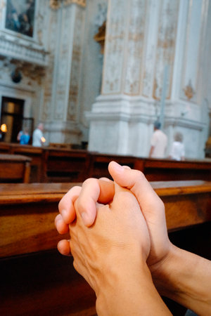 a man sits inside a church with his hands clasped together in prayer resting on a wooden pew while the ornate architecture of the church rises blurred in the backgroundの写真素材