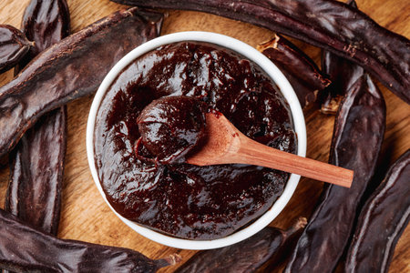 top view of a bowl of dense carob cream, gluten free and natural, with a wooden spoon resting inside, encircled by ripe carob pods on a rustic wooden tableの写真素材