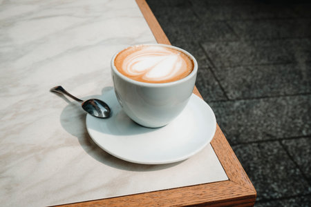 closeup of a macchiato served in a white cup on a saucer with a spoon, showing a heart design in the foam, set on an outdoor cafe table under daylightの写真素材