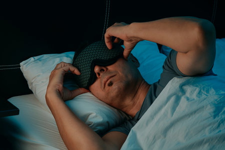 closeup of a man in bed adjusting a patterned sleep mask with both hands in a bedroom, lying on a...