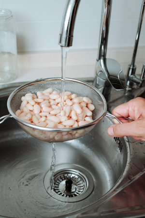 closeup of a man rinsing cooked white beans under running water in a metal strainer, washing them thoroughly at the kitchen sinkの写真素材