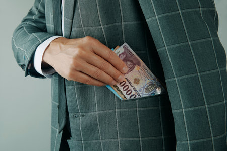 a man wearing a plaid suit putting several Hungarian forint banknotes into his jacket pocket, representing wealth, savings, and financial managementの写真素材