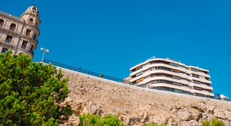 view from below of Balco del Mediterrani in Tarragona, Spain, at the end of Rambla Nova, showing its iron balustrade, modernist architecture on the left, and cliffside stone wall against the skyの写真素材