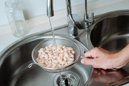 a man holds a metal strainer filled with cooked white beans under a kitchen tap to rinse them, the...