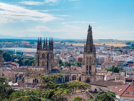 the Gothic towers of Burgos Cathedral, in Spain, rise high over the historic center, surrounded by city rooftops, distant hills, and a bright summer atmosphereの写真素材