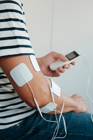 a man holding a portable electrotherapy device while using adhesive pads on his upper arm for muscle stimulation treatment indoorsの写真素材