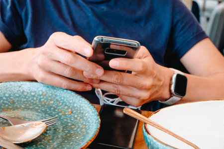 closeup of a man checking his smartphone as it charges from a power bank on a wooden table, with several empty dishes and utensils indicating at the end of a restaurant mealの写真素材