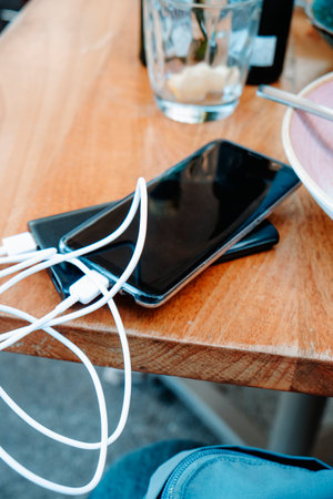 a close view of a smartphone lying on a wooden table with a tangled white cable plugged into a power bank, surrounded by used dishes and a glass after a finished outdoor mealの写真素材