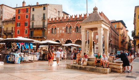 Verona, Italy - June 23, 2025: Tourists rest by the ancient fountain in Piazza delle Erbe, surrounded by colorful buildings and busy market stands beneath the watchful Torre del Gardelloのeditorial素材