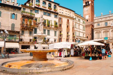 Verona, Italy - June 24, 2025: The fountain of Madonna Verona stands before colorful facades and lively market stalls in Piazza delle Erbe, with the Torre del Gardello rising in the backgroundのeditorial素材
