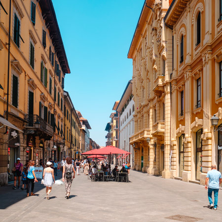 Pisa, Italy - June 26, 2025: A lively view of Corso Italia shows shoppers and pedestrians moving between the warm toned facades and the outdoor cafe tables, shaded by bright red umbrellasのeditorial素材