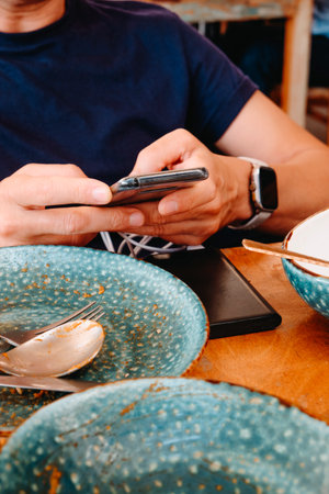 a man seated at an outdoor restaurant table uses his smartphone while it remains plugged into a black power bank, with empty blue plates and cutlery scattered around after lunchの写真素材