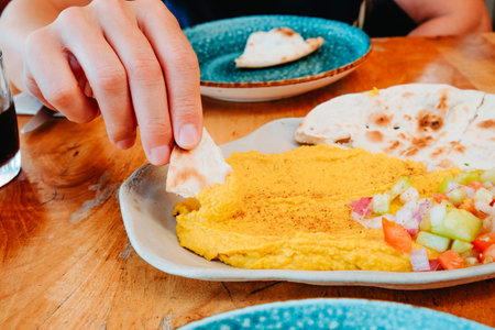 a man lifts naan bread coated with curry hummus from a shared plate on a restaurant table, showing the yellow spread beside fresh chopped vegetables and more naan piecesの写真素材