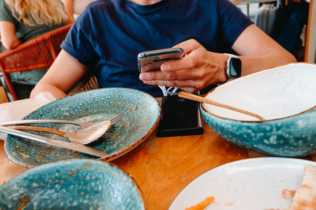 a man holds his smartphone with both hands at a restaurant table while the device stays connected to a power bank, surrounded by empty bowls and cutlery from a recently finished mealの写真素材