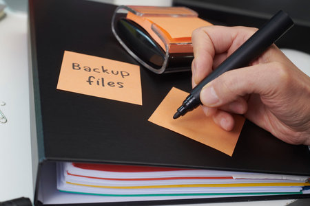 a close-up of a man holding a black marker about to write on a blank orange sticky note on a desk, with another note reading backup files placed on the cover of a ring binderの写真素材