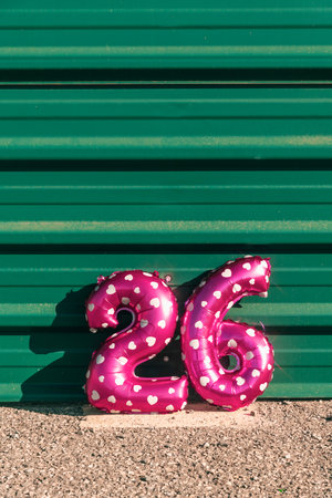 pink foil balloons shaped as the number 26 rest on gravel against a green metal wall, their white...