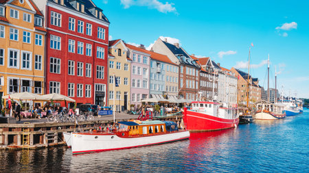 Copenhagen, Denmark - May 12, 2026: Historic boats moored beside the colorful Nyhavn waterfront with restaurant terraces and bright reflections shimmering across the canal