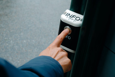 a man presses a silver button on an outdoor public information device mounted on a dark green metal post, with braille lettering above and pavement softly blurred behind