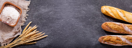 fresh bread with wheat ears and bowl of flour on dark board, top view
