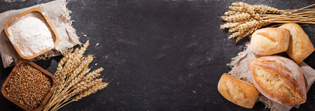 fresh bread with wheat ears and bowl of flour on dark board, top view