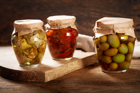 Preserved food. Glass jars of pickled olives, dried tomatoes and artichokes on wooden tableの写真素材