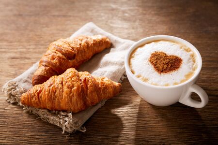 Valentines day.  Cup of cappuccino coffee with drawn heart and fresh croissants on wooden table, top view